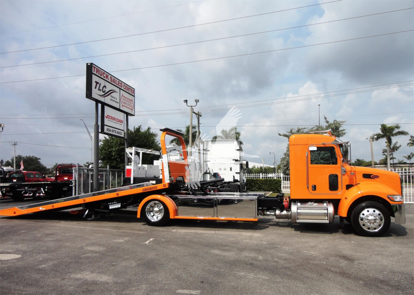 Orange Peterbilt tow truck with a long flatbed, rollback trailer, and white cab; signage on lot in background.