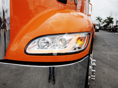 Front left headlight of an orange 2022 Peterbilt 337 rollback truck showing headlight and chrome bumper.