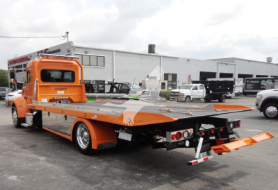 Peterbilt 337 rollback tow truck in orange with flatbed and rear ramps extended. Visible text on cab includes Peterbilt branding; background shows a service lot with buildings and other trucks.