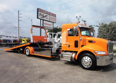 Orange Peterbilt tractor with car hauler rollback trailer in a dealership lot; signs read Truck Sales Center TLC, Jerr-Dan (blue and white sign in background).