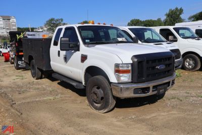 2008 Ford F-350 Super Duty utility truck in a yard, white cab, black front grille, utility body with compartments, amber roof lights, dirt ground.