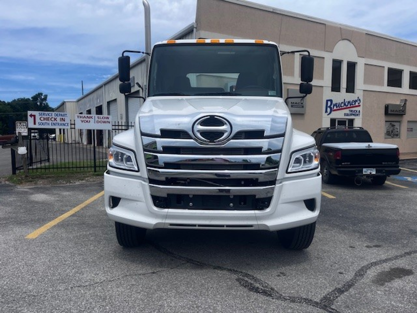 Front view of a white 2026 Hino XL7 cab and chassis truck showing large chrome grille, dual headlights, and emblem; text on building in background.
