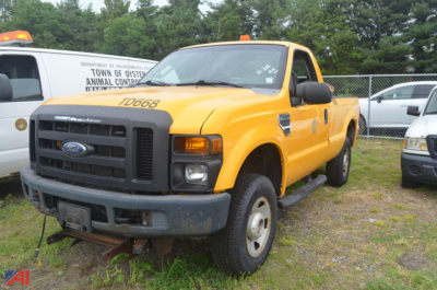 Ford F-250 Super Duty dump truck in yellow viewed from front-left, with black grille, bumper, and side steps; light bar on cab roof; rust at tow hitch area; background shows chain-link fence and other vehicles.