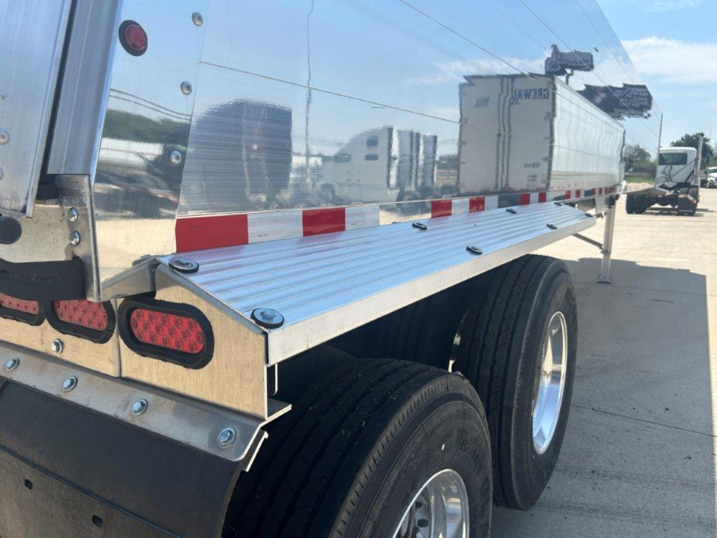 Dump trailer side with tandem axle and aluminum sides showing reflective surface and red/white safety markings.