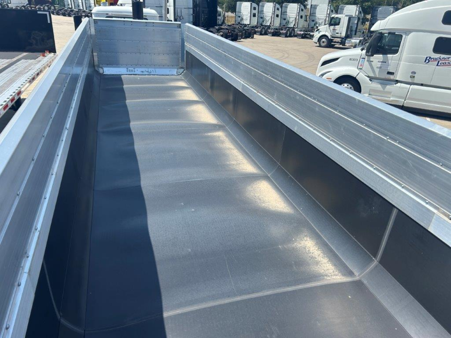 Dump trailer bed interior with smooth metal surface and high sidewalls, silver/gray finish, showing long flat cargo area and rear corner gussets.