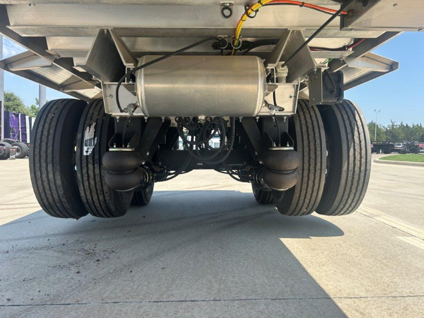 Dump trailer undercarriage showing suspension beams, dual axles, and a central fuel tank/air tank with hoses and wiring visible.