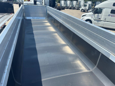 Dump trailer bed interior with smooth metal surface and high sidewalls, silver/gray finish, showing long flat cargo area and rear corner gussets.