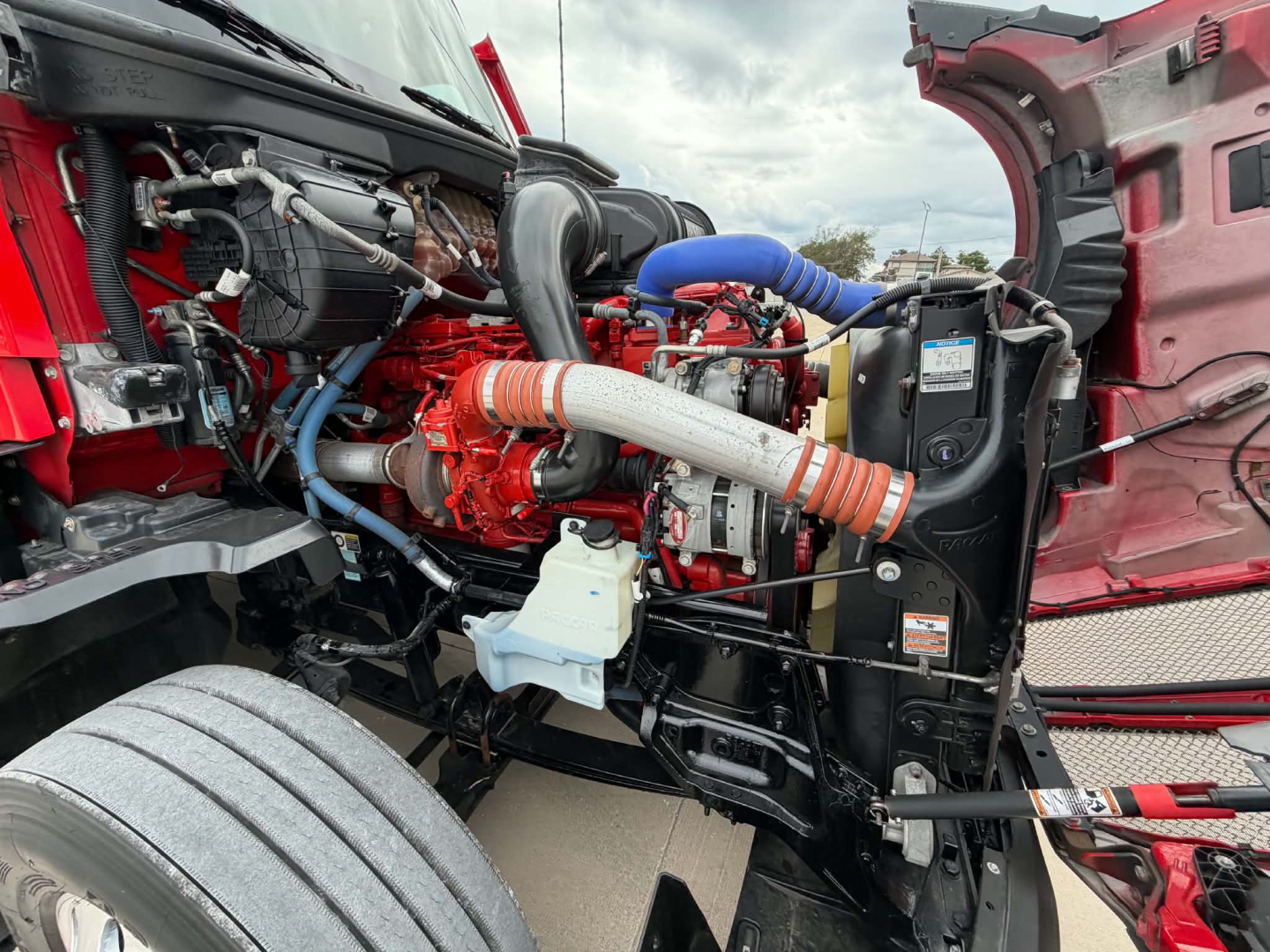 Kenworth T680 engine bay showing red-painted engine with blue and orange hoses, radiator, and various components visible.