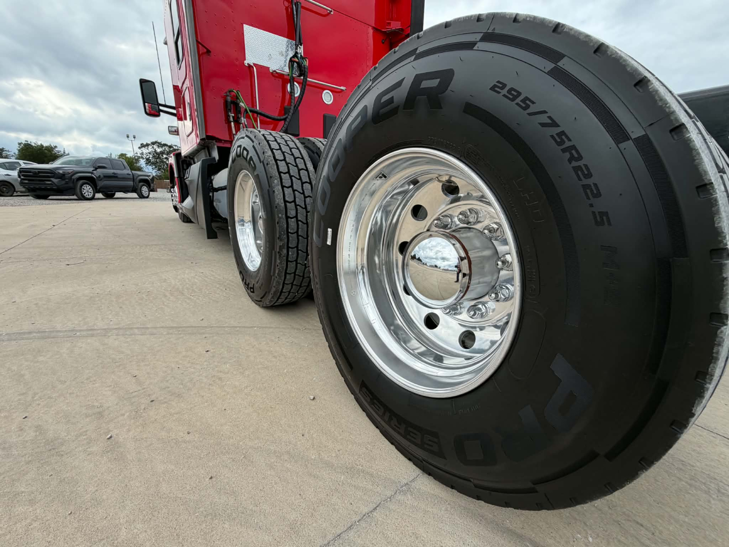 Kenworth T680 truck wheel close-up showing front dual tires with polished chrome rims; tire sidewall reads PIRELLI 295/75R22.5 and other details; cab and red bodywork visible in background.