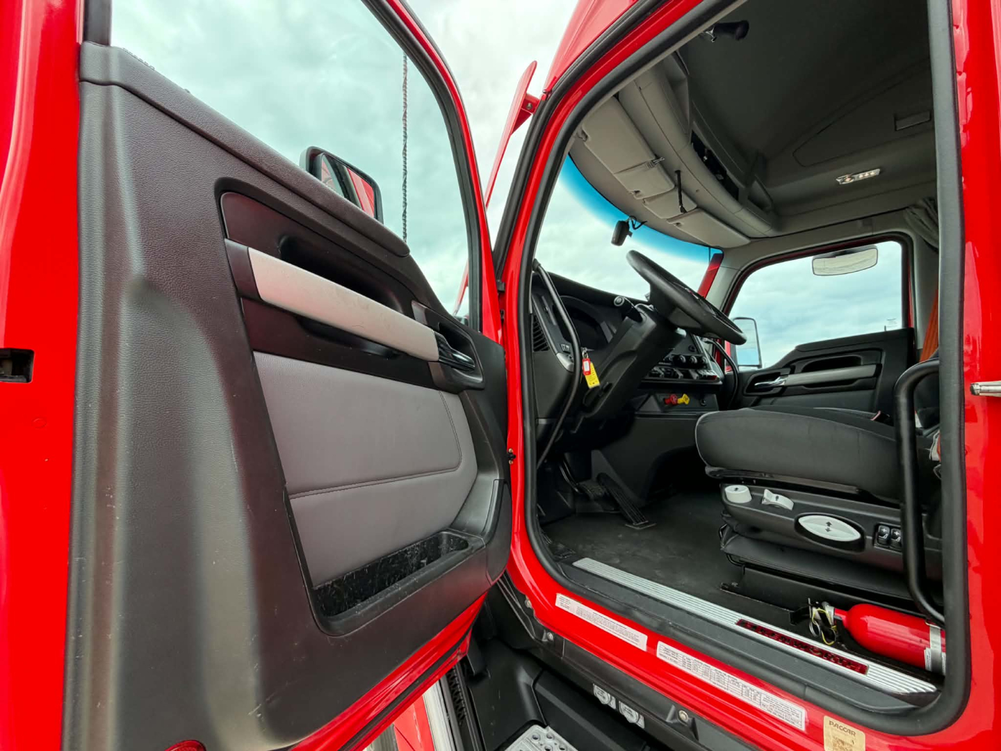 Dashboard and driver seat of a red 2023 Kenworth T680 truck interior with open door; visible steering wheel, driver seat, and entry area.