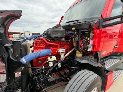 Truck engine bay of a red Kenworth showing a bright red Cummins engine with blue intercooler hose and surrounding components.