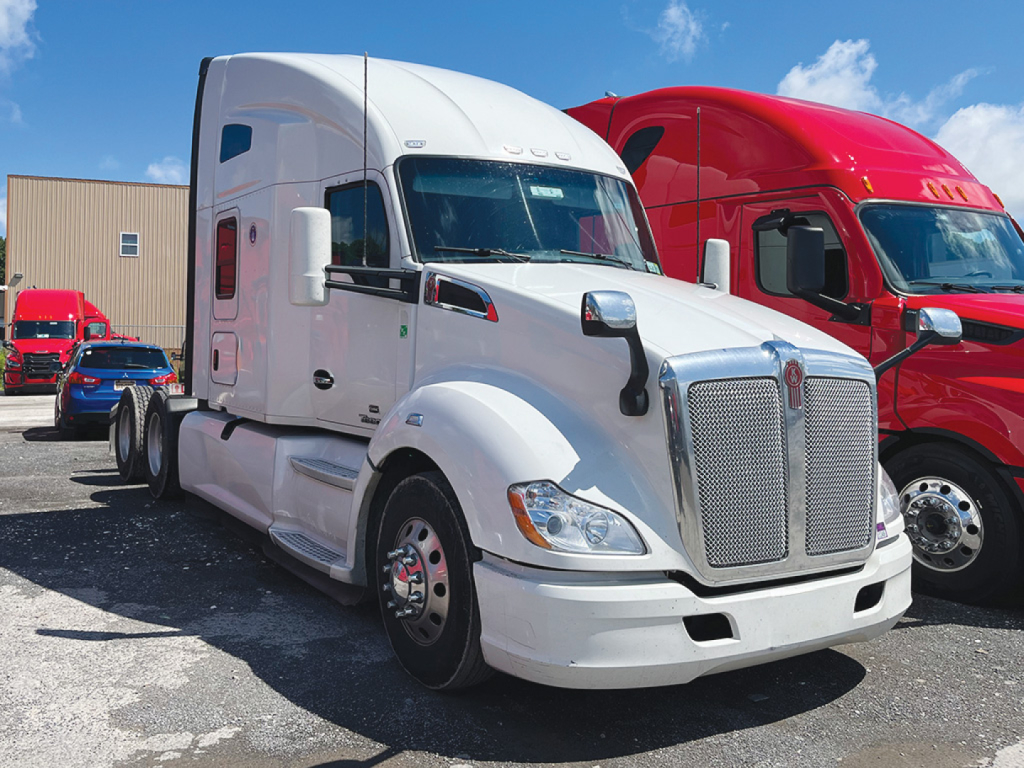 Kenworth T680 tractor-trailer in white with large chrome grille and sleeper cab, parked alongside a red truck.