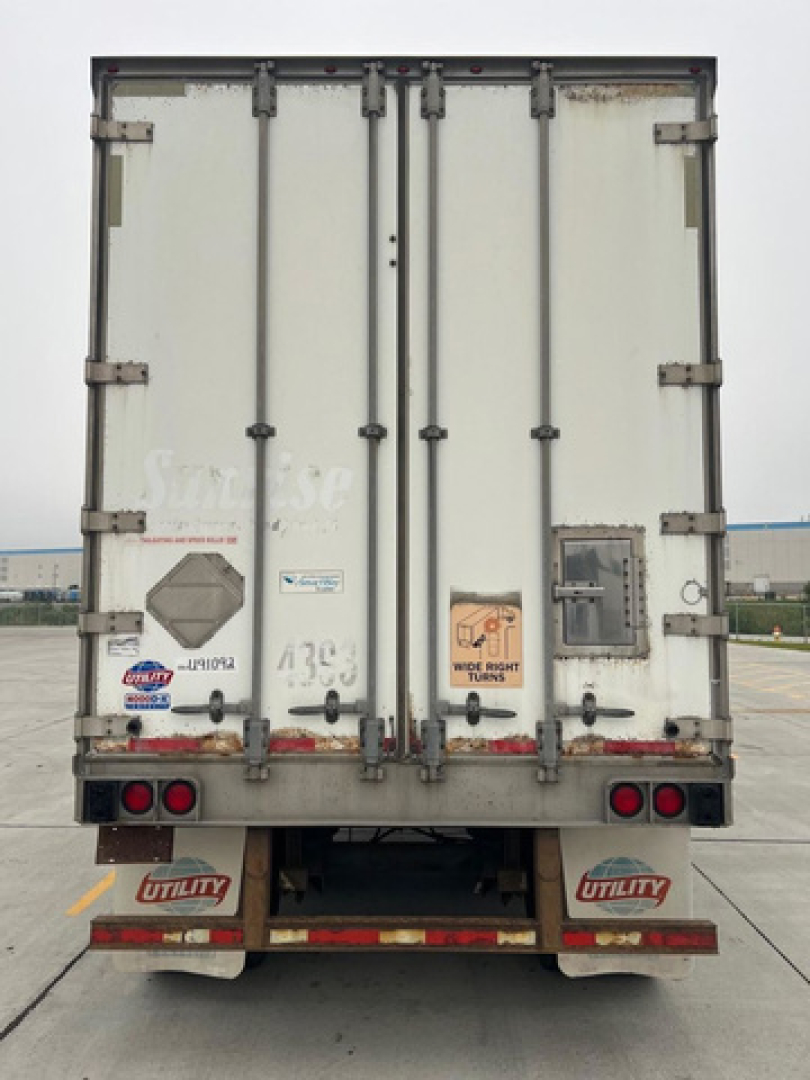 Rear view of a white utility van trailer, doors closed, with red taillights, mud flaps labeled 'UTILITY', and various decals and stickers on the doors.