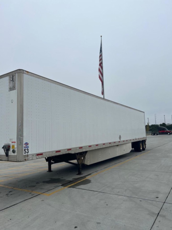 Utility van trailer in a parking lot showing a long white box trailer with landing gear and dual rear wheels; US flag in background; no visible branding.