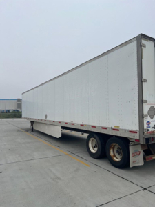 Utility van trailer with tandem rear wheels, long rectangular cargo box, closed roof, and DOT reflective tape along the sides.