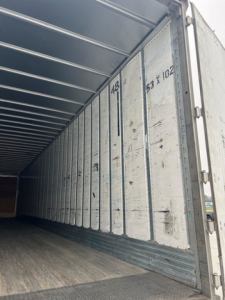 Inside of a 2014 utility van trailer showing metal interior walls with vertical ribs and a wooden floor, door at far end, numbers and wear marks visible