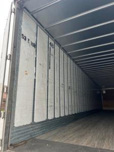 Van trailer interior with vertical corrugated panels and stamped numbers on interior wall; visible text includes '53 & 102' and '98' near the doorway. Left side shows metal skin and wooden floor; light coming from open rear area.
