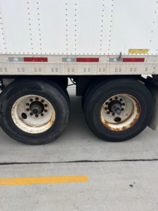 Trailer wheels and side of a utility van trailer showing dual rear axles with black tires and white rims, rusty wheel hubs, and part of the aluminum side panel.