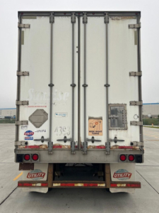 Rear view of a white utility van trailer, doors closed, with red taillights, mud flaps labeled 'UTILITY', and various decals and stickers on the doors.