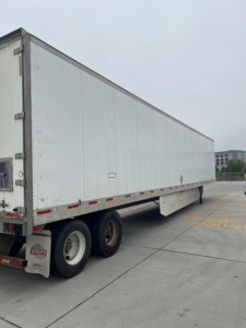 Trailer side view showing a large white utility van trailer with dual rear axles and reflective safety stripes.