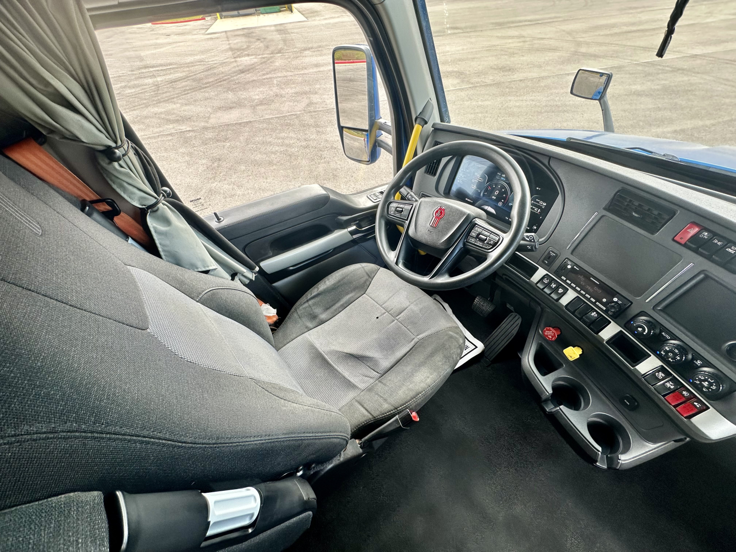 Kenworth T680 cab interior showing driver seat, steering wheel with Kenworth emblem, dashboard with control switches, gray fabric seats, and cup holders.