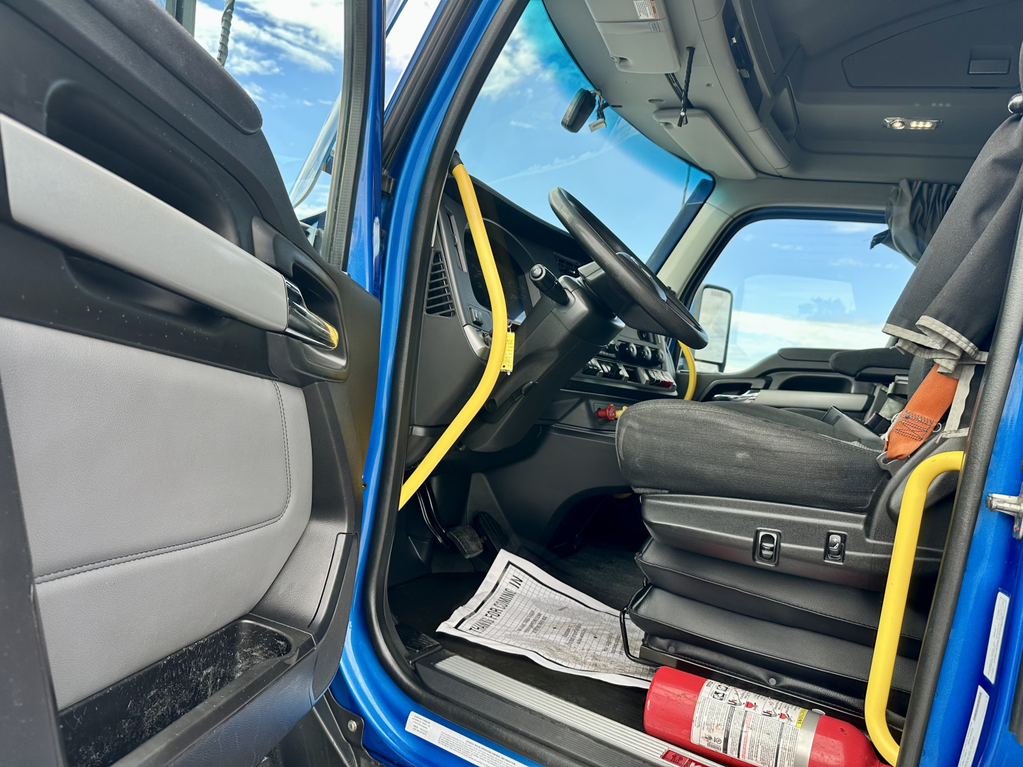 Truck cab interior of a 2023 Kenworth T680 showing blue exterior, driver's seat, steering wheel, yellow grab bars, and dashboard with paperwork on the floor.