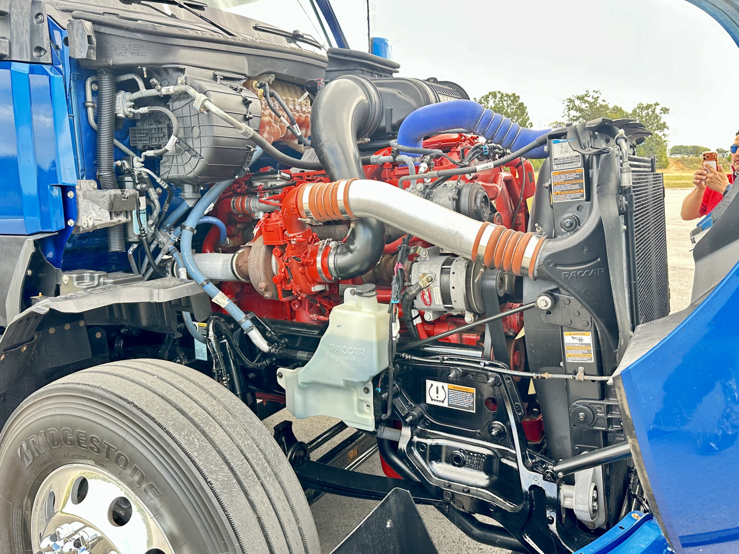 Kenworth T680 engine bay view showing red engine block, blue hoses, orange CV boots, coolant reservoir, and radiator fans.
