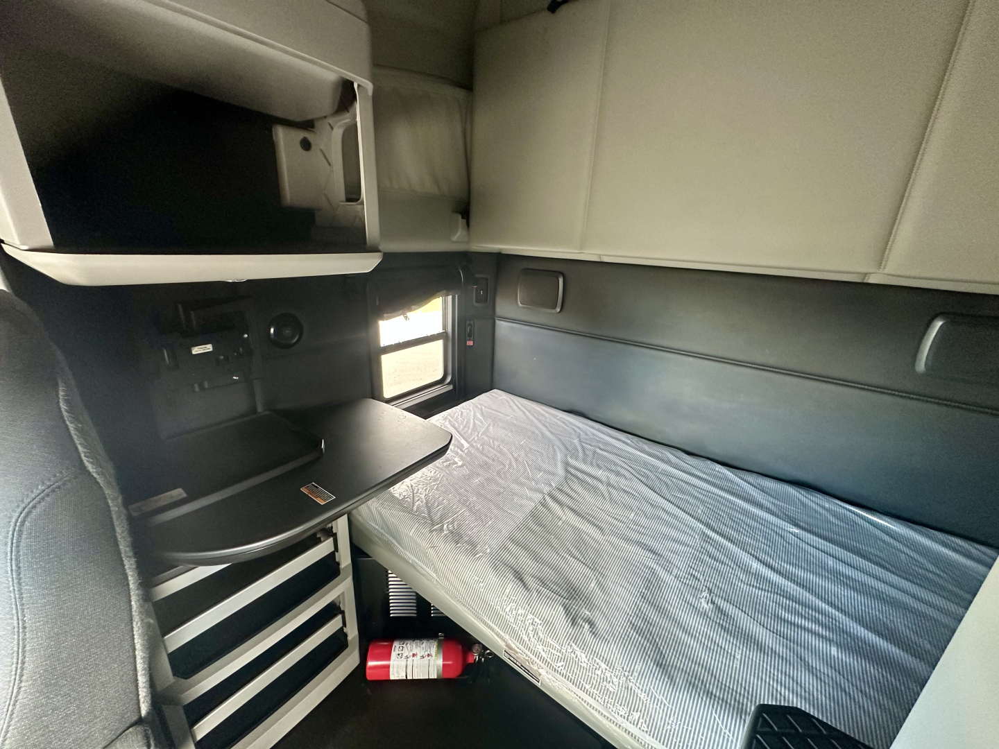 Kenworth T680 sleeper cab interior showing bunk bed with mattress, storage shelves, and small table area.