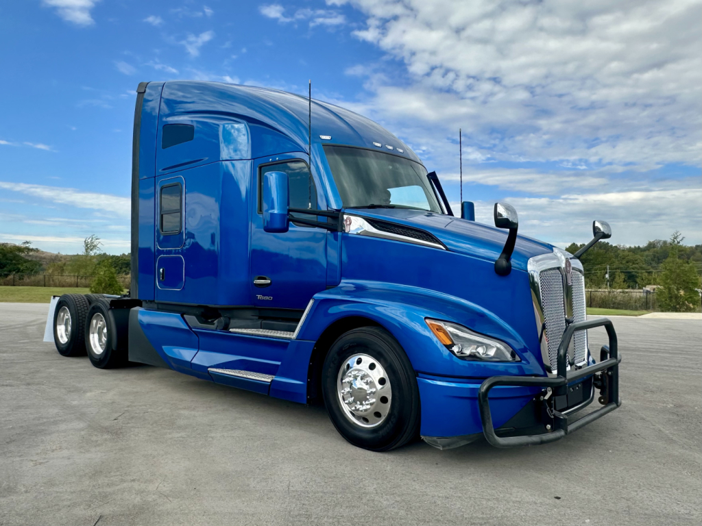 Kenworth T680 blue sleeper cab truck in a parking lot, front three-quarter view, showing chrome grille guard and aerodynamic side mirrors.