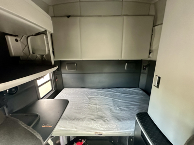 Interior of a Kenworth T680 sleeper cab showing a single bed with mattress, overhead cabinets, and side window; compact work desk area and storage shelves visible.