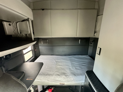 Kenworth T680 sleeper cab interior with a single bed and storage cabinets visible, flat bed mattress covered in plastic, small desk area, and side shelves.