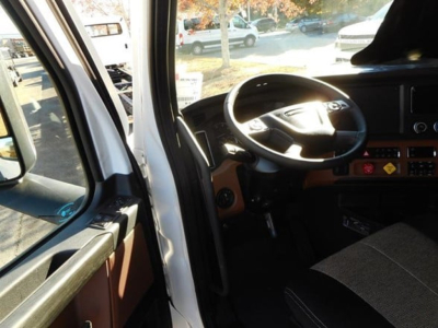 Truck cockpit view from driver side showing steering wheel and dashboard of a Freightliner Cascadia with brown interior trim.