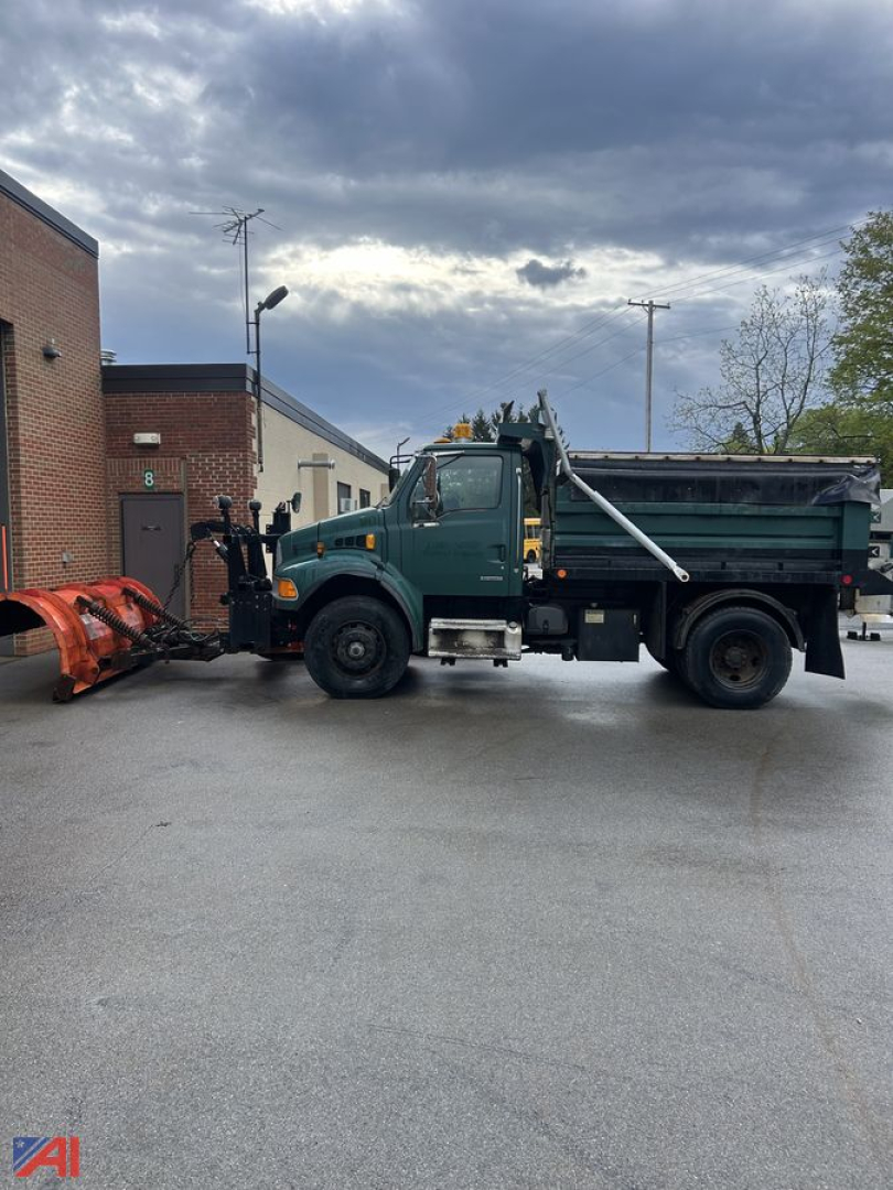 Green Sterling Acterra dump truck with plow attachment and wing, side view parked on pavement near a brick building.