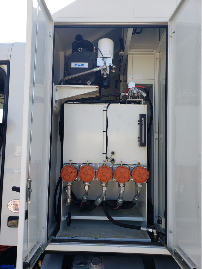 Engine-compartment view of Volvo VHD86B300 trenching truck. Visible components include multiple orange-capped valves/pumps at front of a white tank, hoses, and a compact engine/generator setup inside a vertical compartment.