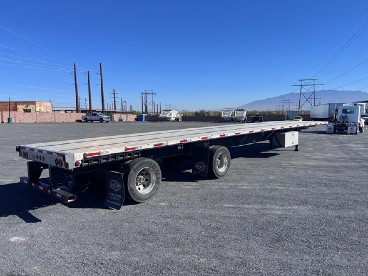 Flatbed trailer with long rectangular deck and dual rear axles, Utility 4000Ae style. Visible text on mudflap and lights; distant vehicles and mountains under clear blue sky.