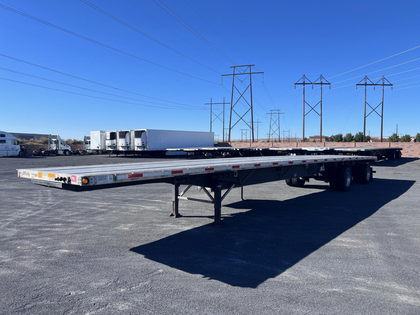 Flatbed trailer showing a long low-profile Utility 4000Ae with dual rear axles and white cab over trucks in distance.