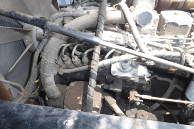 Engine bay view with hoses, belts, and metal components of a used truck engine in a yard setting.