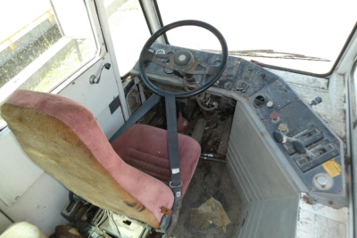 Interior of a used truck cab showing steering wheel, seat, and dashboard with worn controls and pedals; dusty floor and basic instrumentation.
