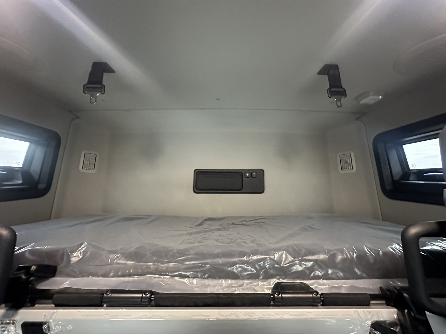 Truck cab interior rear sleeper area with a bed in plastic wrap and overhead mounts.