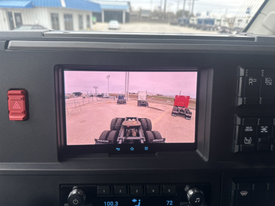 Truck cab dashboard with a rear-view camera display; visible buttons and switches around display. The screen shows road and trailer connections. No specific part numbers readable.
