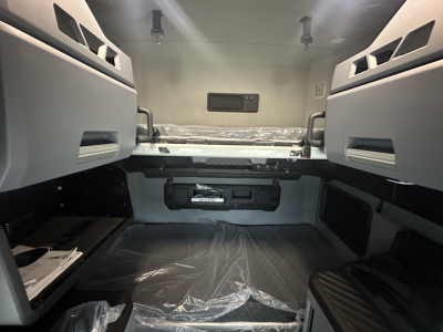 Mack PR64T76H truck sleeper cab interior showing bunk bed area with storage pockets and a center console under window, clean gray surfaces.