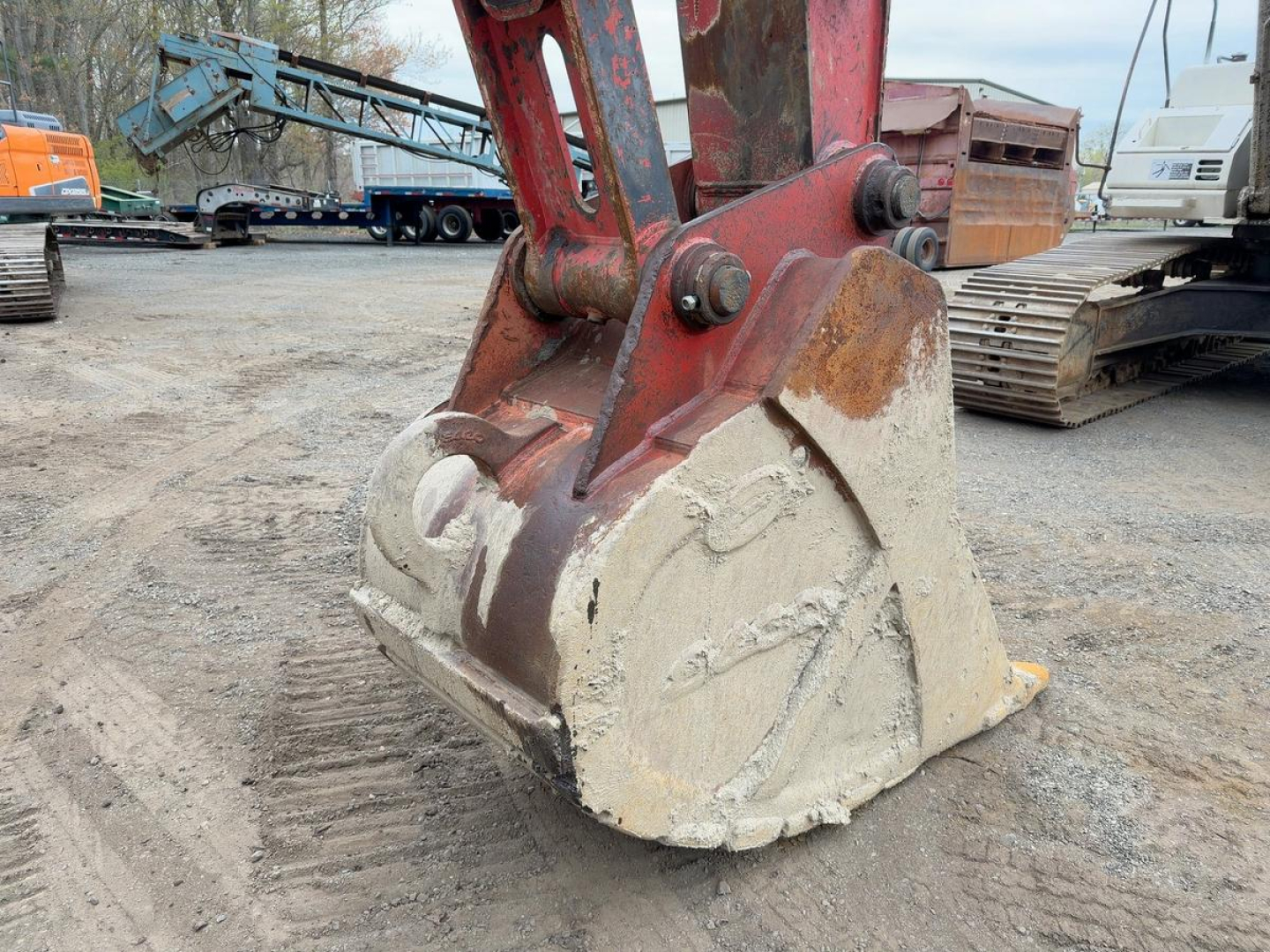 Excavator bucket attachment showing wear marks and hydraulic arm pins in a workyard, with other heavy equipment in the background.
