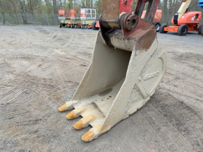 Excavator bucket with teeth on the ground, hydraulic arm attachment visible in a yard with other construction equipment in the background.