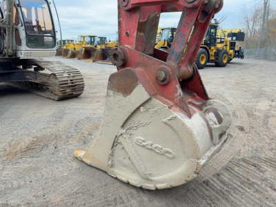 Excavator bucket attachment with Link-Belt logo and hydraulic arm in a yard showing other equipment in background.