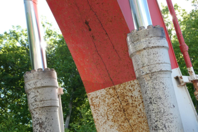 Excavator undercarriage components with hydraulic struts and rust-patterned wear, front view showing two large cylindrical hydraulic cylinders and red boom.