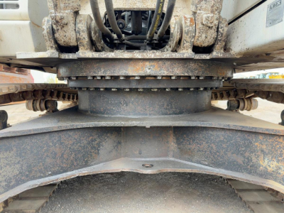 Excavator undercarriage showing large rotating swing bearing and tracks with hydraulic hoses and rust-prone surfaces.