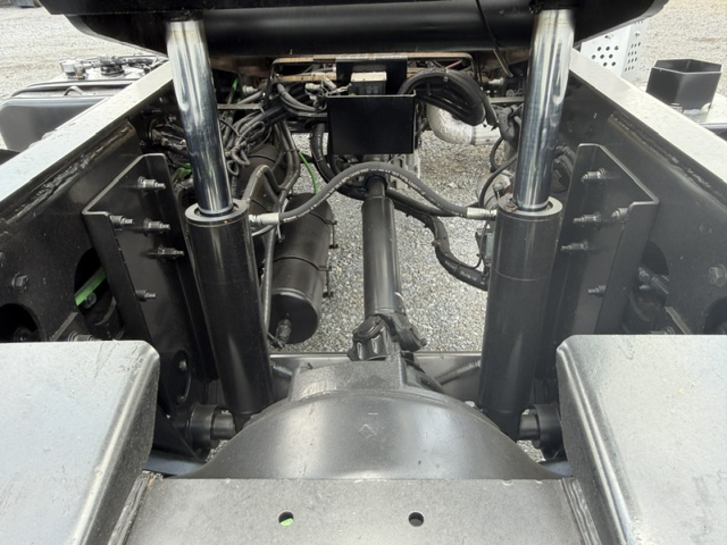 Truck chassis and hydraulic system viewed from rear, showing steel suspension struts, hoses, and hydraulic components beneath cab, with drivetrain housing in center.
