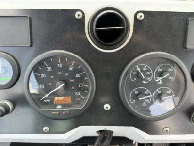 Dashboard of a Capacity Sabre truck showing two round gauges: left gauge with speedometer up to 120 MPH and odometer display; right gauge with multiple small dials.  Center circular vent above, screws at corners, plain black panel.