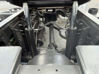Truck chassis and hydraulic system viewed from rear, showing steel suspension struts, hoses, and hydraulic components beneath cab, with drivetrain housing in center.