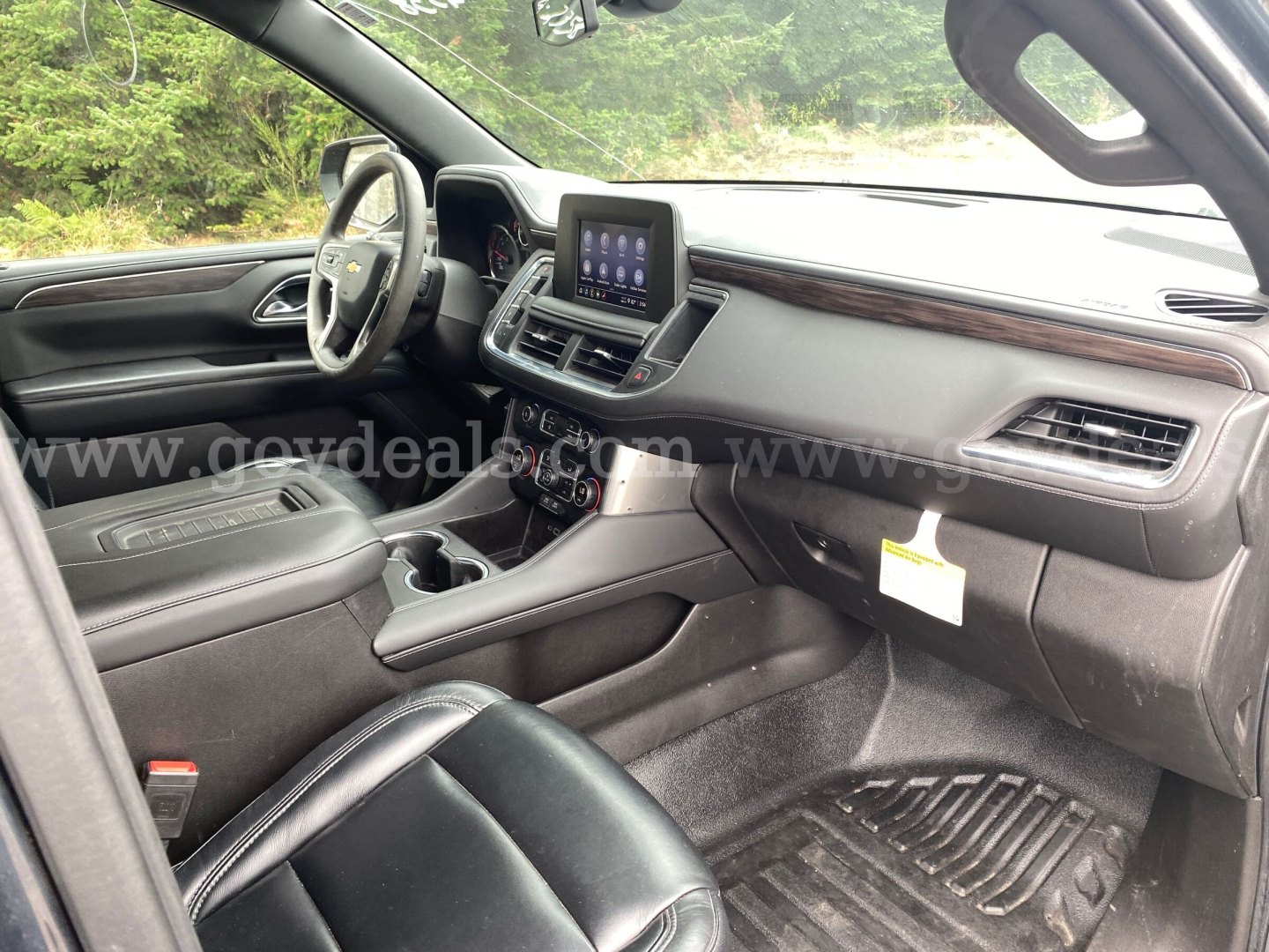 Interior of 2021 Chevrolet Suburban SUV front cabin showing steering wheel, center touchscreen, dashboard, and front seats; black leather seats with wood trim; gear selector area and climate controls visible.